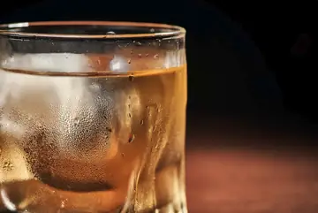 A glass containing a drink and an ice cude. on a table top. There's a dark background, it it probably evening time.