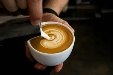 A coffee in a white porcelain cup with frothed milk being poured into it by a barista